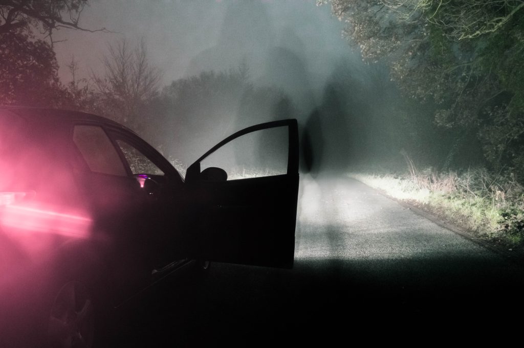 The image shows an abandoned car surrounded by fog facing a ghostly apparition.